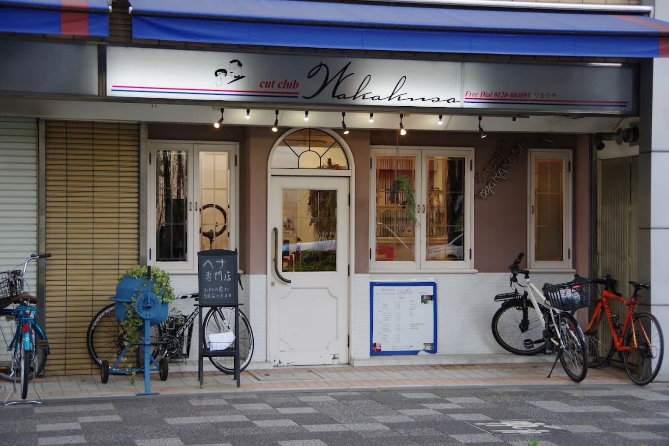 Le magasin a une enseigne qui dit « cut club Wakana » au-dessus d’une double porte blanche, et offre des services de raccourci et de henné. Pris en sandwich entre les fenêtres et le parking à vélos, il y a des supports à vélos bleus, des plantes d’intérieur, des menus et des panneaux près de l’entrée.