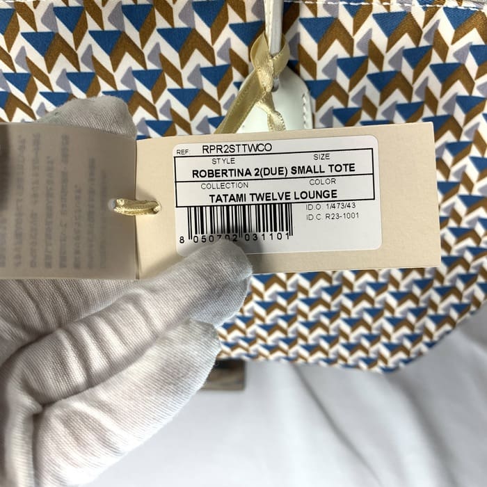 La main gantée tient l’étiquette du produit « ROBERTINA 2 (DUE) SMALL TOTE » sur le tissu à motif tatami avec un motif géométrique en bleu, marron et blanc, qui est le style signature de la collection de petits fourre-tout de ROBERTA PIERI.
