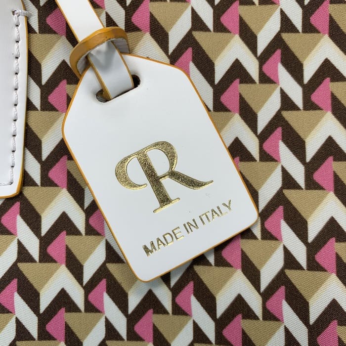 Gros plan d’une étiquette de bagage Roberta Pieri blanche avec l’inscription « R » en or et la mention « MADE IN ITALY » attachée à un mini sac de sport aux motifs géométriques marron, beige et rose.