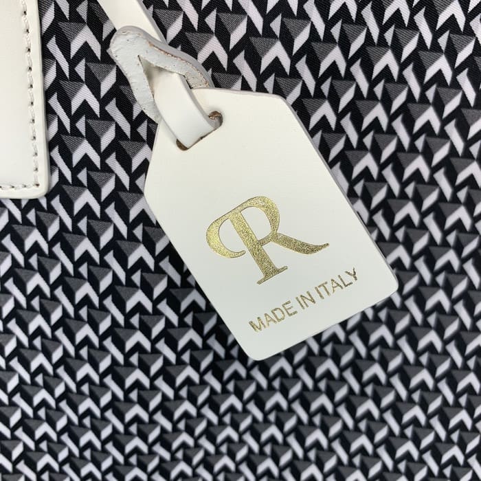 Une étiquette ROBERTA PIERI en cuir blanc avec les lettres « R » en or et les mots « MADE IN ITALY » est suspendue au fond d’un tissu géométrique noir et blanc.