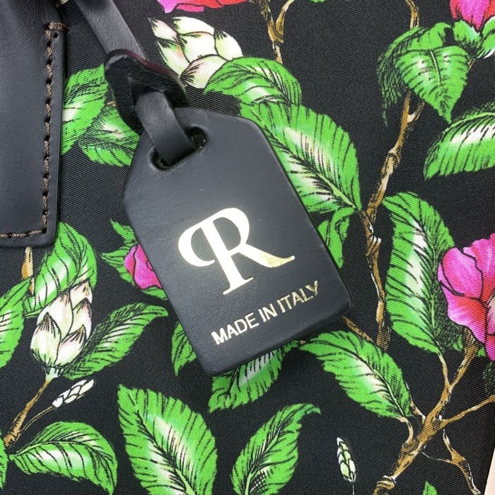 L’étiquette de bagage noire ROBERTA PIERI avec le « R » doré et le texte italien est suspendue à un tissu floral avec des feuilles vertes et des fleurs roses, complétant le mini sac de sport avec une élégance italienne intemporelle.