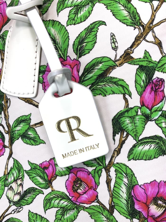 Gros plan du mini sac de sport ROBERTA PIERI. pédoncules à feuilles vertes et fleurs roses ; L’étiquette blanche attachée au sac à 2 voies comporte un logo « R » doré et les mots « MADE IN ITALY ».